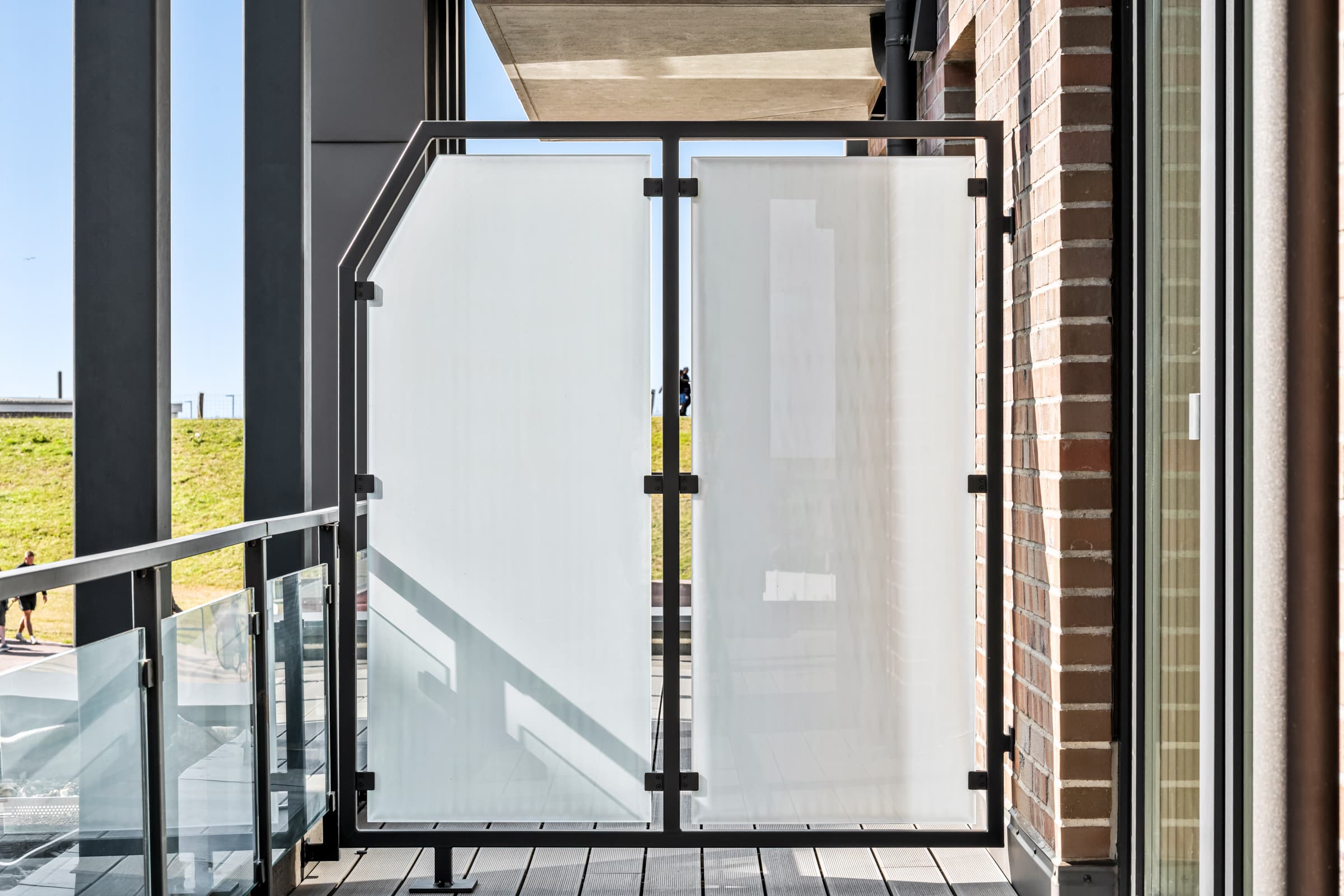Balkon-Trennwand aus schwarzem Stahlrahmen mit zwei satinierten Glasfeldern, dahinter Klinkerfassade und Deich