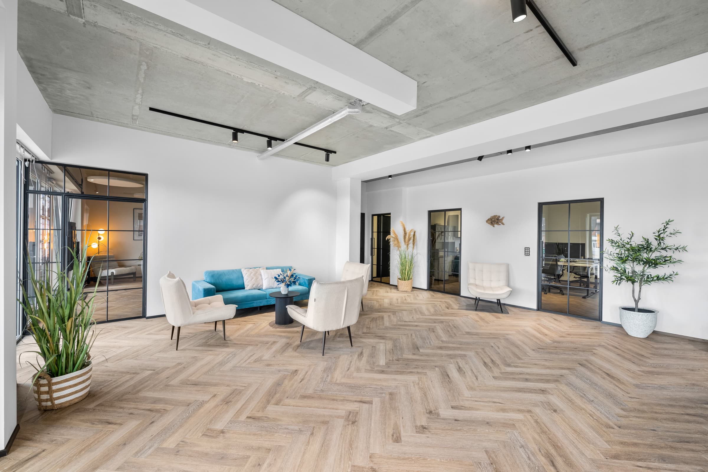 Büro-Lounge mit türkisem Sofa, Fischgrätparkett und raumhohen Stahl-Glas-Trennwänden unter Sichtbetondecke