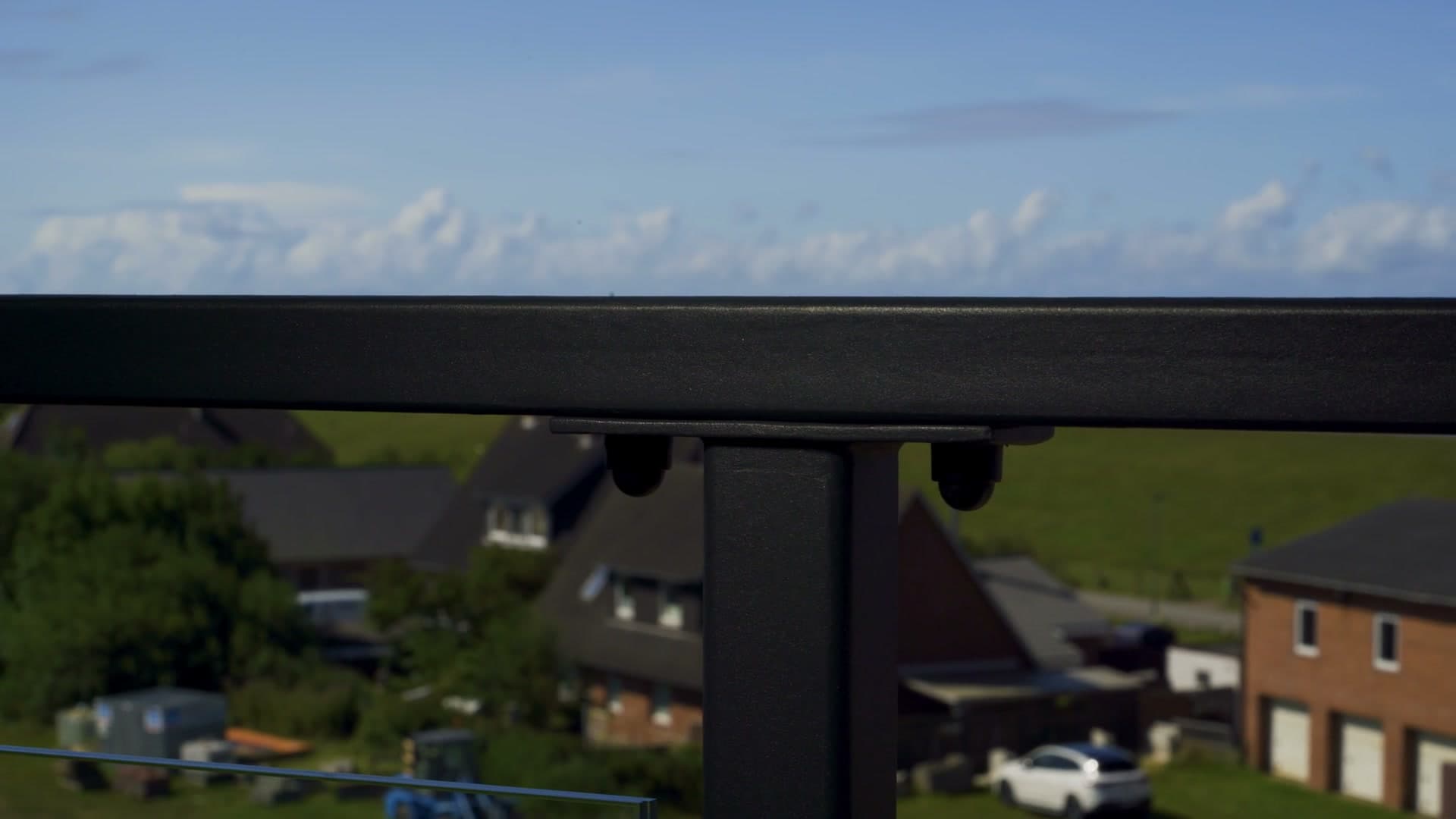 Kamerafahrt entlang des Handlaufs einer Balkon-Glasbrüstung, dahinter Häuser und Marschwiese